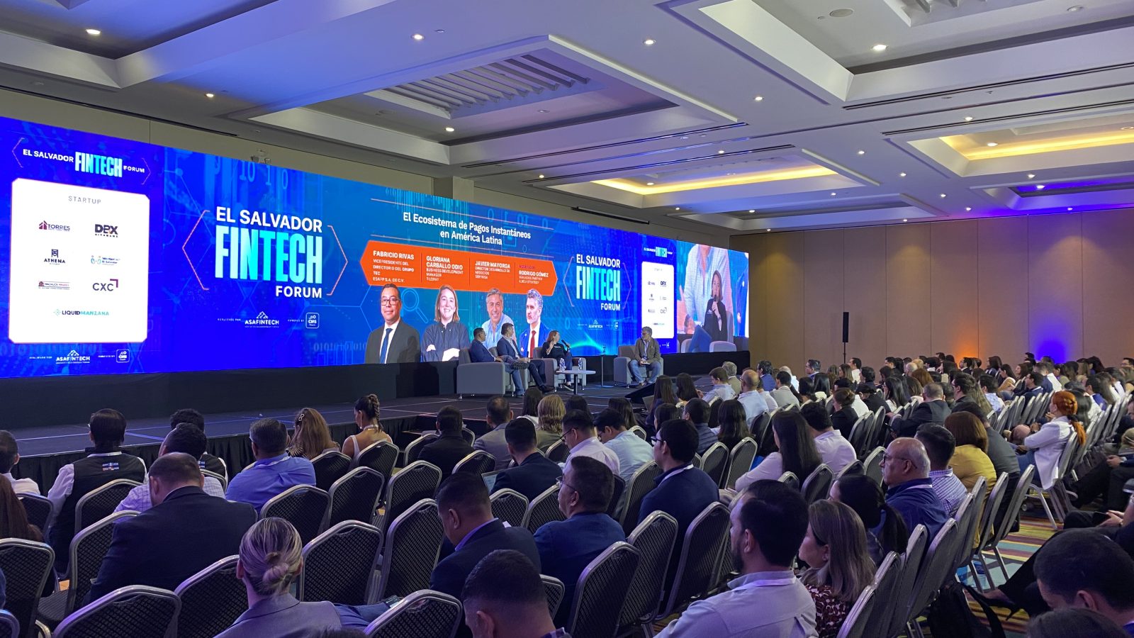 Panel en el escenario principal de El Salvador Fintech Forum, con pantalla de patrocinadores y audiencia llena.